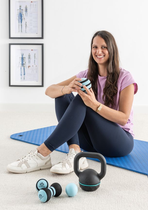 Friederike Döll sitzt sympathisch lächelnd auf einer Trainingsmatte und hält einen kleinen Fitnessball in den Händen. Vor ihr liegen zusätzlich eine Kettlebell und eine kleine Fitnesshantel.